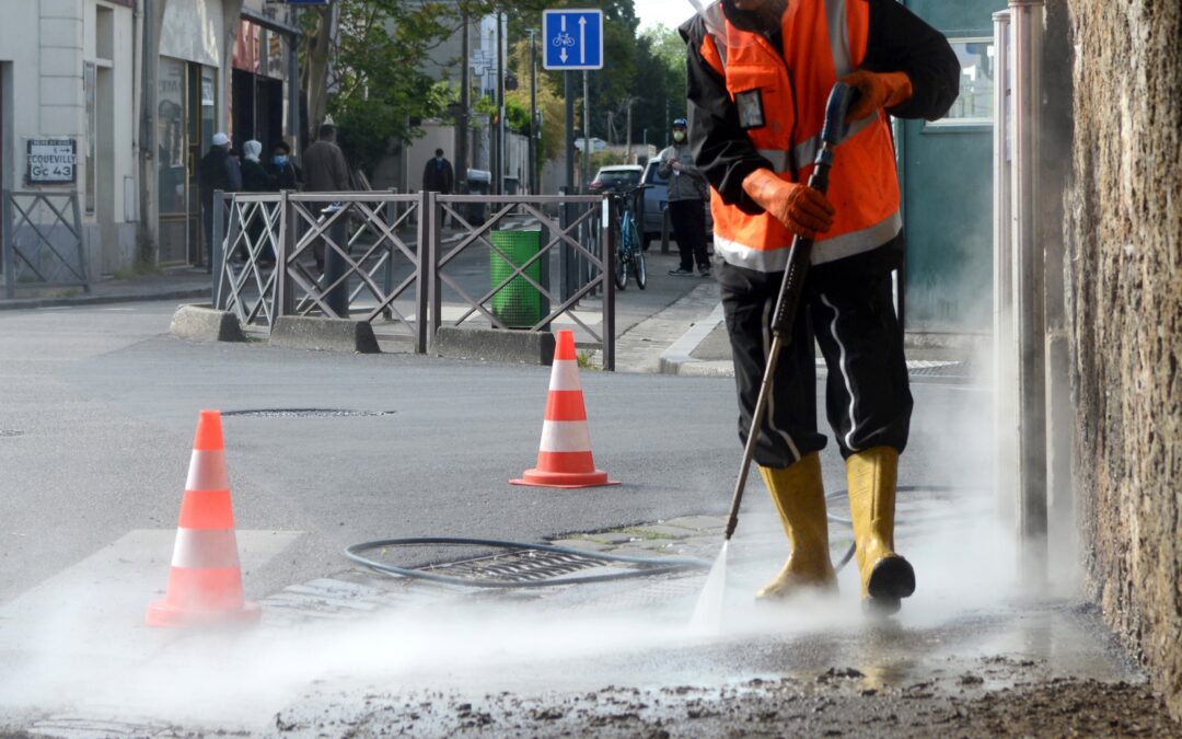 A worker wearing safety gear pressure-washing a sidewalk next to traffic cones on a city street