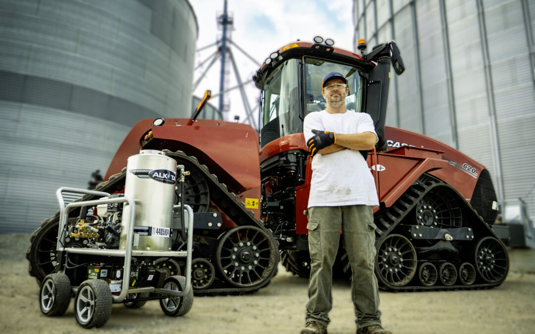 A worker standing confidently in front of a large tracked tractor with high-pressure cleaning equipment beside him on an industrial site.